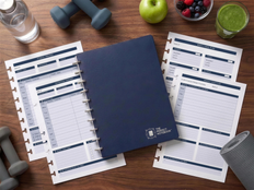 The Wellness Kit from The Perfect Notebook: navy B5 disc-bound notebook with fitness tracker, meal plan and wellness refill pages, dumbbells, water and fruit on a wooden desk